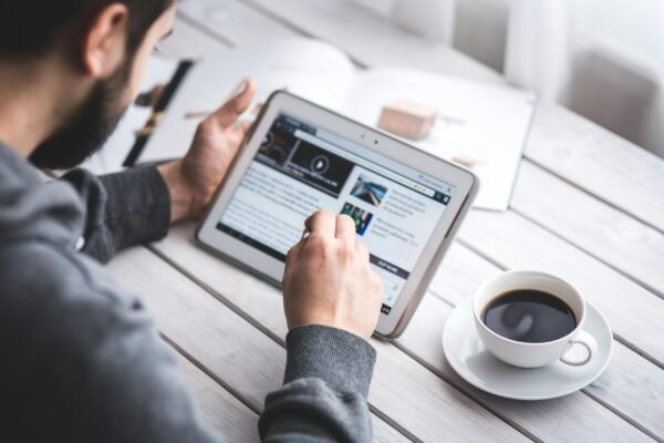 Alcance real: como transformar um simples post em matéria de portal A man uses a tablet to browse news while enjoying a cup of coffee, stylus in hand.